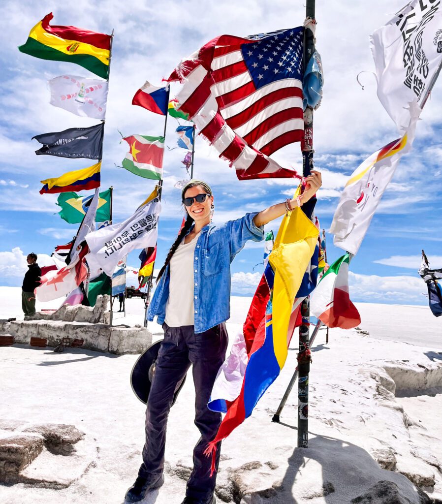 a tourist who saved money for travel poses next to many flags set up on a salt flat.