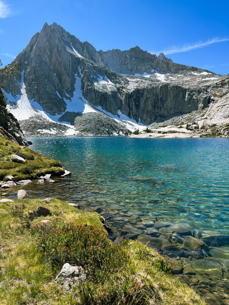 The bright turquoise Hungry Packer Lake, contrasted by a green meadow next to it and craggy mountain peaks in the background.