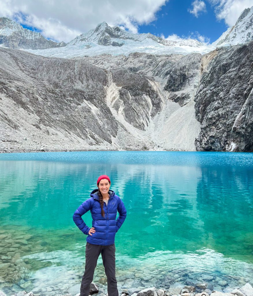 a day hiker with cold-weather hiking gear from a hiking packing list stands in front of a turquoise alpine lake and craggy, snowy mountains.