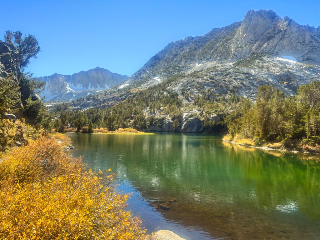 a pristine alpine lake on a california hike surrounded by golden foliage, green meadows, and tall mountains.