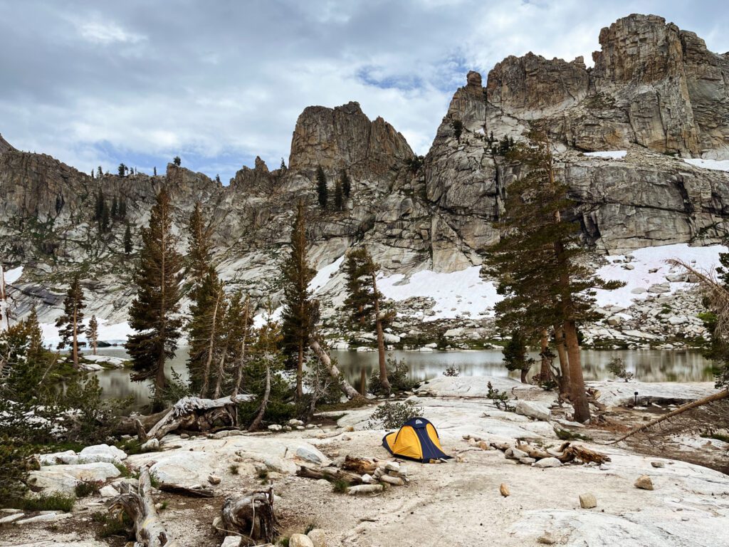 A tent pitched near the rocky lakeshore of pear lake, with tall snowy peaks in the background.