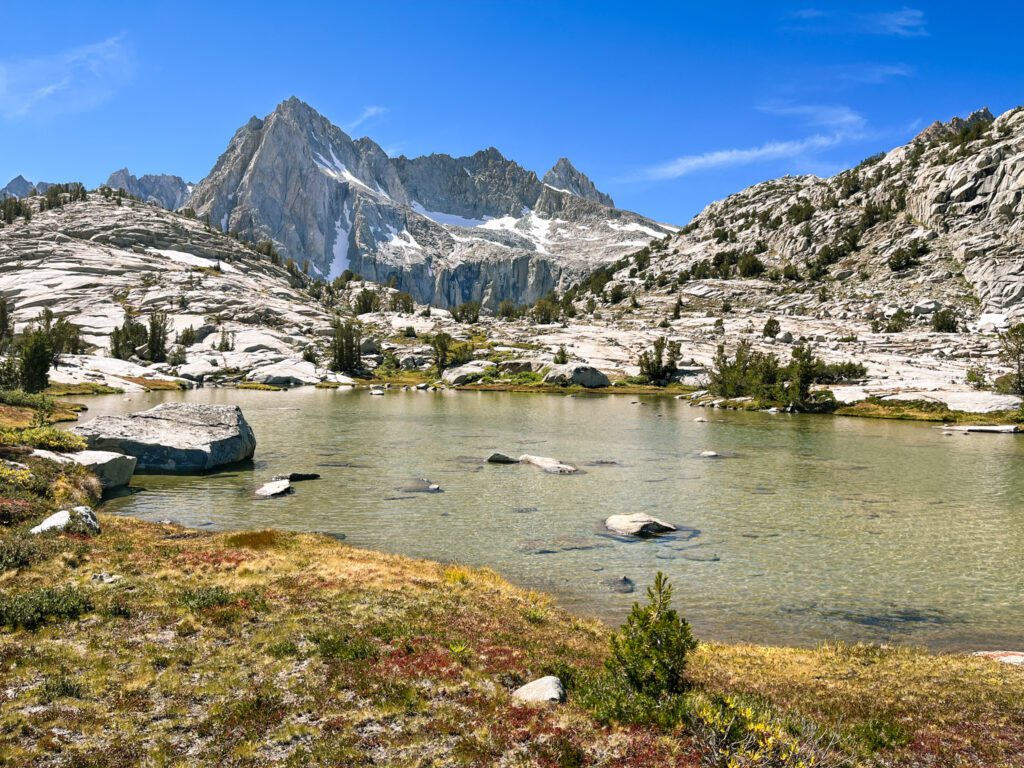 a shallow, light teal alpine lake surrounded by a colorful meadow and craggy mountains.